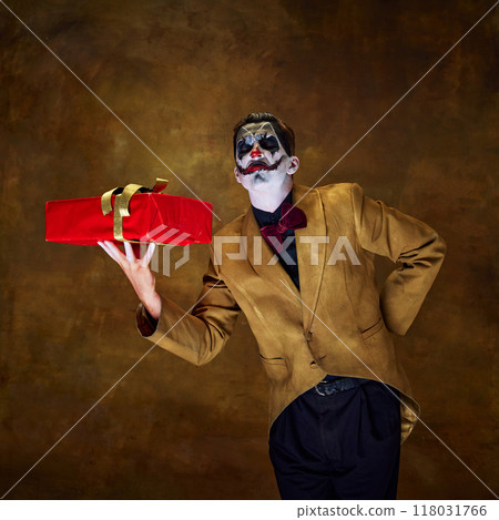 Proudly holding red gift, clown grins mischievously, as if presenting something dangerous against vintage dark backdrop. 118031766
