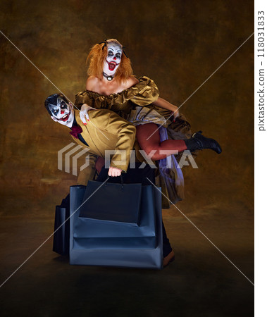 Woman in creepy clown make-up, riding on partner's back, both in vintage outfits and posed dramatically against dark background. 118031833
