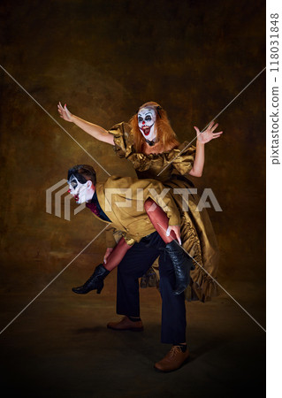 Woman in clown make-up, sitting on partner's back, both dressed in vintage attire, striking playful balance against vintage background. 118031848