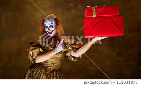 Creepy clown woman, dressed golden dress grinning widely as presents red gift box posing against vintage style background. 118031875