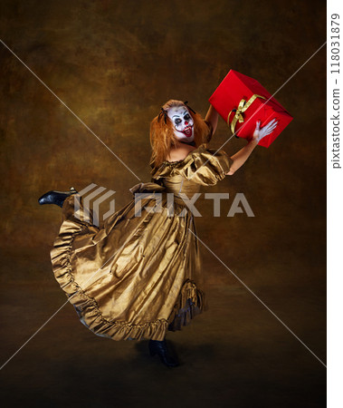 Frightening clown woman dressed in old-fashioned gown, lift leg up while holding red gift box against vintage style background. 118031879