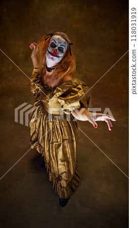 Frightening clown woman in golden gown, raising her hands with wild expression, posing dramatically against vintage background. 118031919