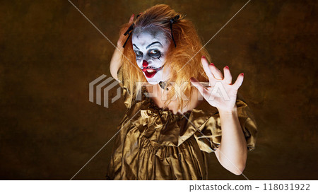 Frightening clown woman in golden gown, raising her hands with wild expression, posing dramatically against vintage background. 118031922