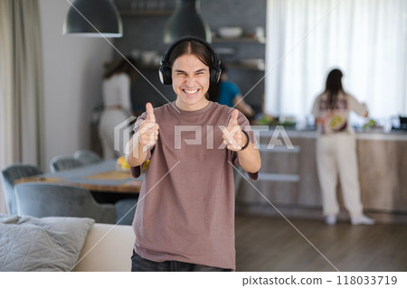 Cute teenage boy dance during listening music in living room. Background of thee females preparing food in the kitchen. Thumbs up on both hands Cute teenage boy dance during listening music in living room. Background of thee females preparing food in the kitchen. Thumbs up on both hands 118033719