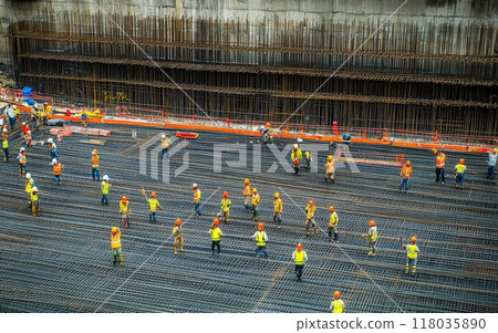 Aerial view of massive construction site with numerous workers in yellow and orange safety gear, working on reinforced steel framework for a large concrete structure 118035890