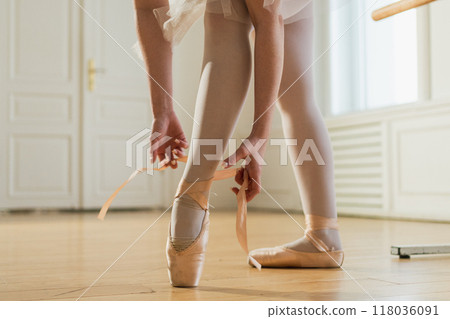 Ballerina puts on pointe shoes on leg in dance class. Classical ballet dancer woman in dance studio. Ballerina putting on tying classical pointe shoes for dance training. Ballet school concept Ballerina puts on pointe shoes on leg in dance class. Classical ballet dancer woman in dance studio. Ballerina putting on tying classical pointe shoes for dance training. Ballet school concept 118036091