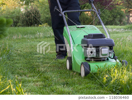 Man pushing a green lawn mower across a lush garden lawn on a sunny day. Man pushing a green lawn mower across a lush garden lawn on a sunny day. 118036460