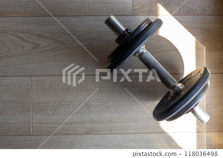 A single dumbbell resting on a wooden floor with shadows, highlighting a fitness setup in natural light. A single dumbbell resting on a wooden floor with shadows, highlighting a fitness setup in natural light. 118036498
