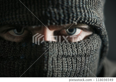 Intense close-up of a person's eyes peering through a knitted balaclava mask in low lighting. Intense close-up of a person's eyes peering through a knitted balaclava mask in low lighting. 118036549