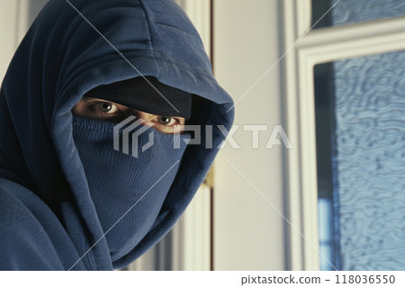 Close-up of a masked intruder in a blue hoodie, staring intently at the camera. Close-up of a masked intruder in a blue hoodie, staring intently at the camera. 118036550
