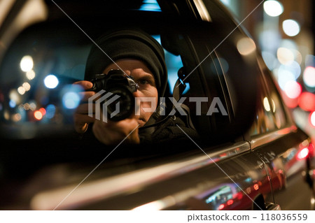 Hooded photographer capturing images from inside a car during nighttime, with city lights in the background. Hooded photographer capturing images from inside a car during nighttime, with city lights in the background. 118036559