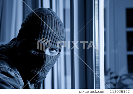A masked man peers through a window at night, with a dark, moody atmosphere. A masked man peers through a window at night, with a dark, moody atmosphere. 118036562