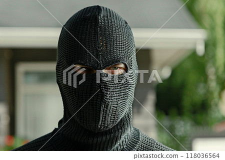 A masked criminal standing in front of a house, wearing a black balaclava and a textured outfit. 118036564