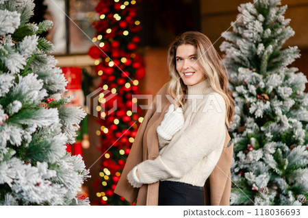 portrait of a happy woman in a coat and white mittens posing against the backdrop of Christmas decorations, the mood of the approaching holiday portrait of a happy woman in a coat and white mittens posing against the backdrop of Christmas decorations, the mood of the approaching holiday 118036693