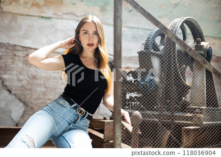 Woman in black t-shirt and jeans standing near wire fencing in abandoned factory 118036904