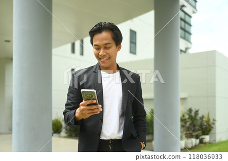 Smiling young asian man dressed in a business suit using mobile phone outside office building 118036933