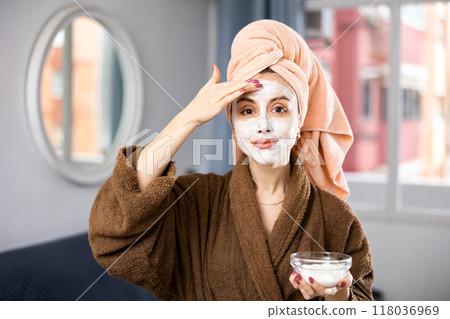 Woman in bathrobe smoothing face with cream and looking at camera 118036969