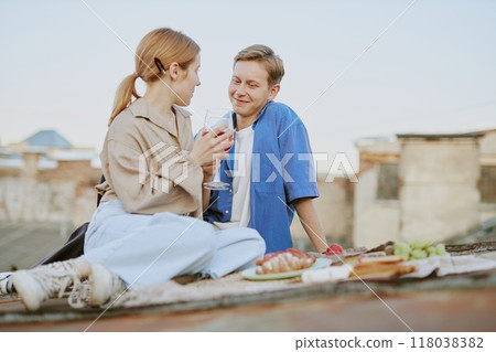 Young couple sitting on a rooftop enjoying picnic and each others company while holding drinks and smiling surrounded by food and scenic view of cityscape Young couple sitting on a rooftop enjoying picnic and each others company while holding drinks and smiling surrounded by food and scenic view of cityscape 118038382