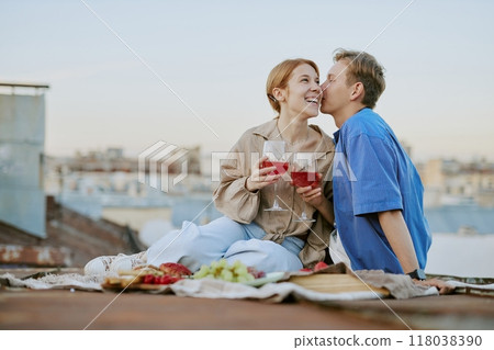 Couple sharing an intimate moment while sitting on a rooftop, holding glasses of wine and smiling, nearby romantic setup with fruits and blanket, cityscape in background Couple sharing an intimate moment while sitting on a rooftop, holding glasses of wine and smiling, nearby romantic setup with fruits and blanket, cityscape in background 118038390