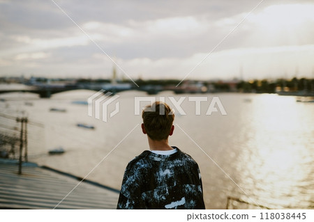 Individual gazing at city skyline from elevated platform near water. Sunlight softly illuminating the scene while boats dot the river in background Individual gazing at city skyline from elevated platform near water. Sunlight softly illuminating the scene while boats dot the river in background 118038445