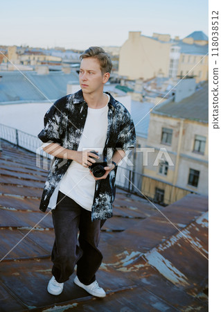 Young man standing on rooftop holding camera, with cityscape in background looking thoughtful. Casual apparel and urban surroundings with sunset lighting atmosphere Young man standing on rooftop holding camera, with cityscape in background looking thoughtful. Casual apparel and urban surroundings with sunset lighting atmosphere 118038512