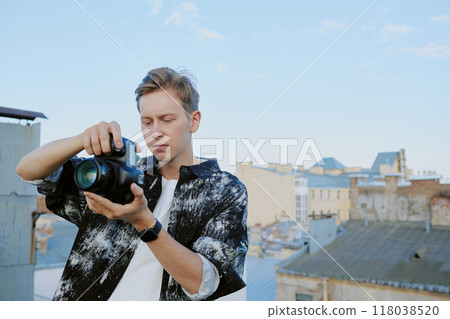 Young Caucasian man adjusting camera settings on urban rooftop during daylight hours. He is deeply focused on the device, with city buildings visible in the background 118038520