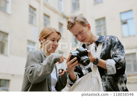 Young individuals examining camera settings in outdoor cityscape, surrounded by buildings, one holding professional camera while other points at screen, showcasing interest in photography 118038546