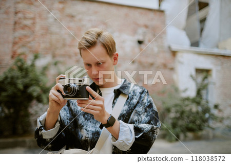 Young Caucasian man adjusting vintage camera outdoors, preparing for shot in urban environment with greenery and rustic walls behind 118038572
