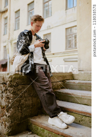 Young man holding camera while sitting on outdoor stairs of an old building, deeply focused on both camera and surroundings, casually dressed with urban backdrop providing context 118038578
