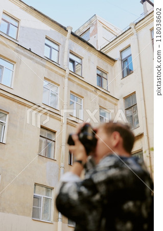 Man photographing exterior of tall old building with vintage camera leaving a blurred effect in foreground adding artistic touch to urban scene 118038602