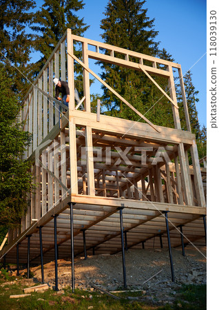 Joiner building wooden, two-story frame house near the forest. Bearded man in glasses hammering nail with protective helmet on. The concept of modern ecological construction. 118039130