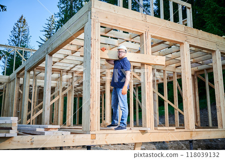 Carpenter constructs wooden-framed house near forest. Man holds large truss on the shoulder while dressed in work clothes and helmet. An idea of modern and eco-friendly construction. Carpenter constructs wooden-framed house near forest. Man holds large truss on the shoulder while dressed in work clothes and helmet. An idea of modern and eco-friendly construction. 118039132