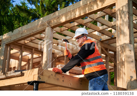 Carpenter constructing wooden frame house near forest. Bearded man with glasses hammering nails into structure, wearing protective helmet, construction vest. Concept of modern ecological construction. 118039138