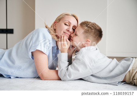 Little son showing love to his mom. Young mother and her kid lying on white carpet and spending time together at home. Lovely afternoon with family. Little son showing love to his mom. Young mother and her kid lying on white carpet and spending time together at home. Lovely afternoon with family. 118039157