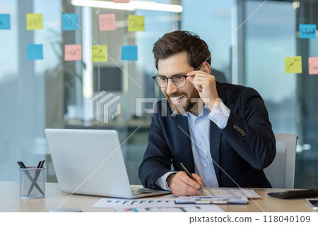 Confident businessman wearing glasses working on laptop in office setting with sticky notes. Displays positive energy, focus, success, and modern workplace environment. 118040109