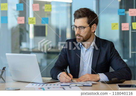 Businessman concentrating on work at desk with laptop, taking notes in modern office environment. Businessman concentrating on work at desk with laptop, taking notes in modern office environment. 118040116