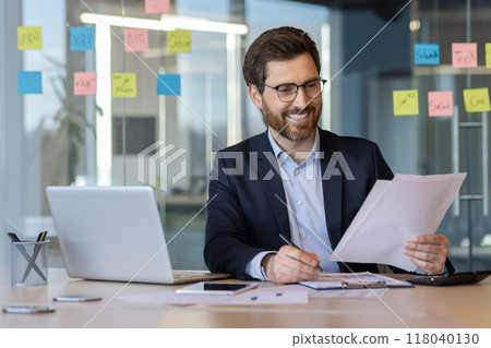 Cheerful businessman examining paperwork at office desk using laptop and smartphone for efficient work. Cheerful businessman examining paperwork at office desk using laptop and smartphone for efficient work. 118040130
