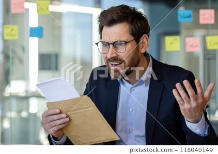 Businessman in glasses expressing confusion while reading letter in office. Represents stress, surprise, uncertainty in workplace. Ideal for concepts of communication, reaction, challenge 118040149
