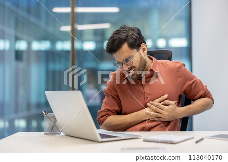 Businessman at desk holds chest in discomfort, displaying symptoms of health issue during work. Laptop on desk suggests busy office environment. 118040170
