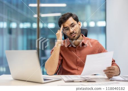 Businessman feeling stressed while reviewing documents at office desk. Man appears confused and frustrated, surrounded by laptop and paperwork, conveying workplace stress and challenges in corporate 118040182
