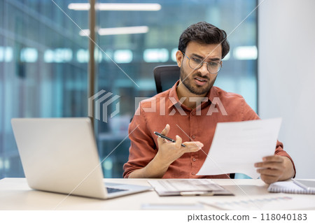 Businessman examining document while seated at office desk with laptop open. Wearing glasses and appearing concerned. Workplace setting with focus on critical thinking and decision-making. Businessman examining document while seated at office desk with laptop open. Wearing glasses and appearing concerned. Workplace setting with focus on critical thinking and decision-making. 118040183