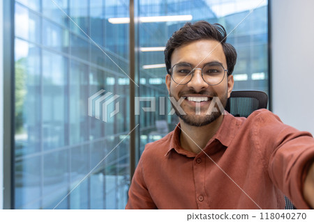 Confident young businessman smiling for selfie using smartphone in modern office environment. Displays success, positivity, and professionalism. Perfect for promoting positive workplace culture 118040270