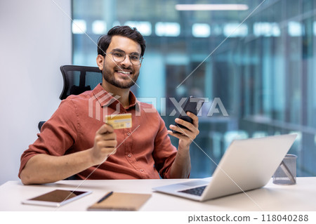 Smiling man in office holding phone and credit card at desk with laptop. Concept of online banking, digital payment, work, and technology. Confident and happy professional connecting digitally. 118040288