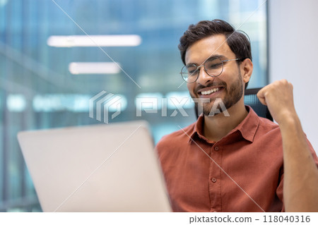 Young professional expressing joy and success while looking at laptop. Celebratory fist pump and smile convey satisfaction, achievement, and positive energy in modern office environment. Young professional expressing joy and success while looking at laptop. Celebratory fist pump and smile convey satisfaction, achievement, and positive energy in modern office environment. 118040316