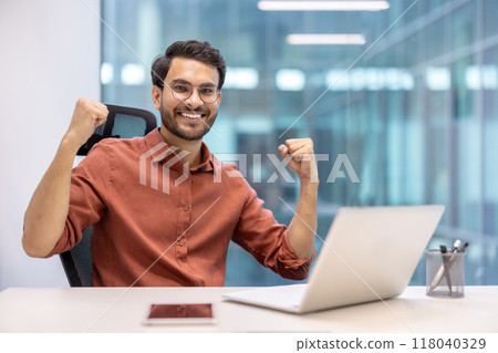 Young professional with glasses celebrating success at desk with laptop and smartphone. His joyful expression conveys achievement and productivity in modern office setting. 118040329