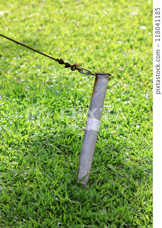Rusty metal pole or iron stake is buried in the ground and is firmly fixed to the rope. Close up photo of an iron tent peg with rope stuck into the ground at a camping site. Rusty metal pole or iron stake is buried in the ground and is firmly fixed to the rope. Close up photo of an iron tent peg with rope stuck into the ground at a camping site. 118041185