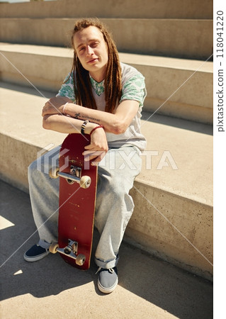 Full shot of teenage guy with tattoo on arm holding skateboard while posing on concrete bench looking at camera on sunny day outside Full shot of teenage guy with tattoo on arm holding skateboard while posing on concrete bench looking at camera on sunny day outside 118041220