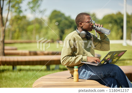 Side view of young African American male student on bench studying using laptop and drinking water to stay hydrated on sunny day, casual outdoor learning scene, copy space 118041237
