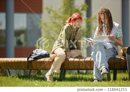 Smiling female student with male friend on bench reading notes in notebook together making learning fun while studying outdoors on sunny day, copy space 118041253