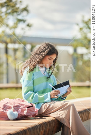 Curly teenage girl on bench writing in notebook doing homework while afternoon sunlight casting warm glow on her hair in park, casual outdoor learning scene Curly teenage girl on bench writing in notebook doing homework while afternoon sunlight casting warm glow on her hair in park, casual outdoor learning scene 118041272